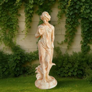 Graceful lady sculpture in a soft marble finish, holding a flower and draped in flowing attire, displayed outdoors against a vine-covered wall and greenery.