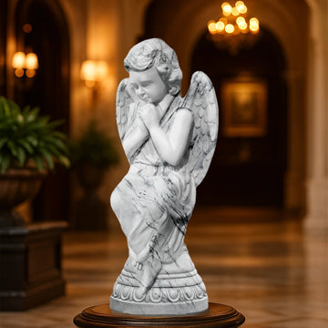 Graceful praying angel sculpture in a white marble finish with fine detailing, displayed indoors on a pedestal in a luxury hall with chandelier lighting.
