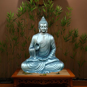 Serene meditating Buddha sculpture in a metallic silver-blue finish, seated in lotus posture on a carved wooden pedestal, with bamboo plants in the background for a peaceful setting.
