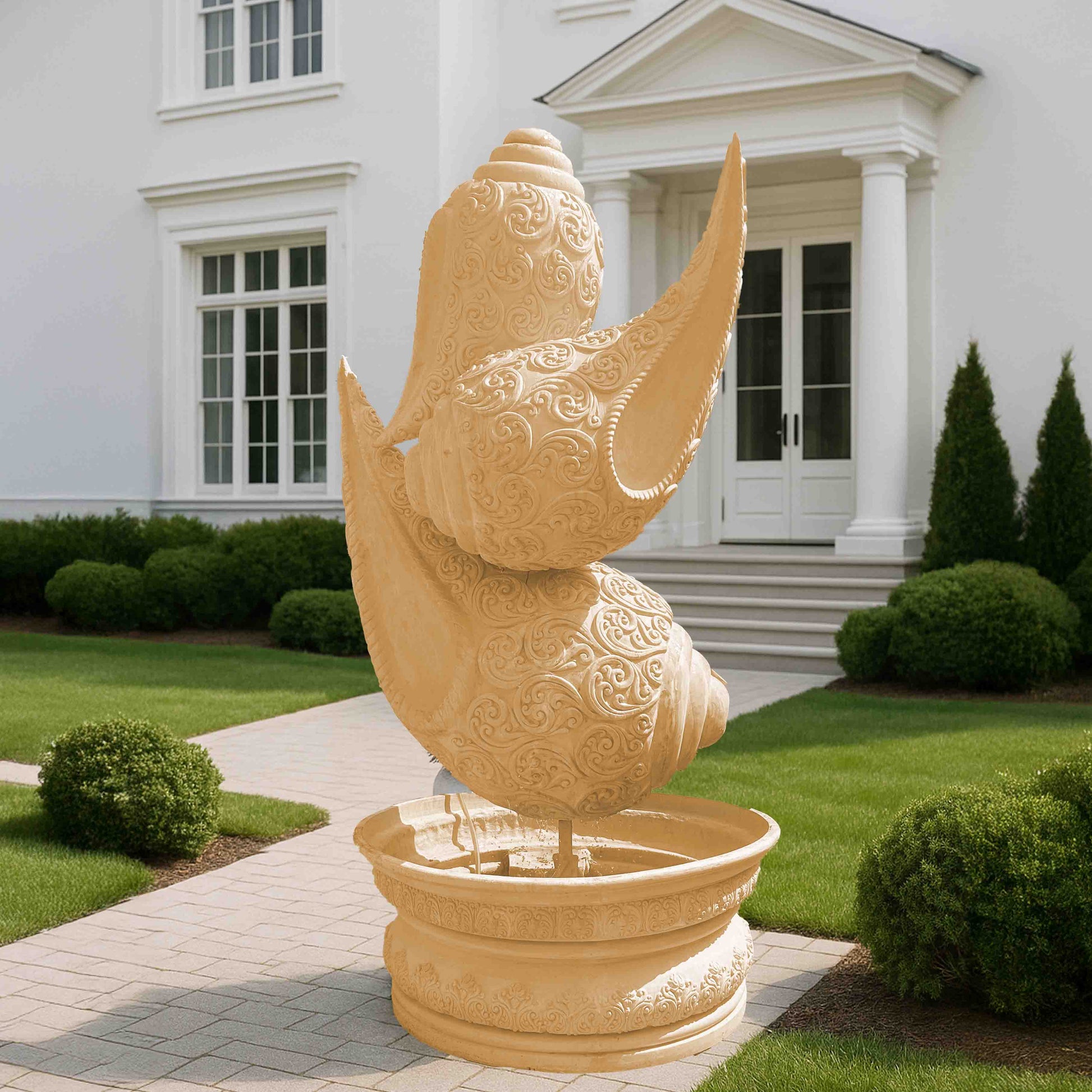 Intricately carved beige stone-finish shankh (conch shell) fountain sculpture with ornate detailing, placed on a decorative pedestal basin in an elegant garden entrance setting.