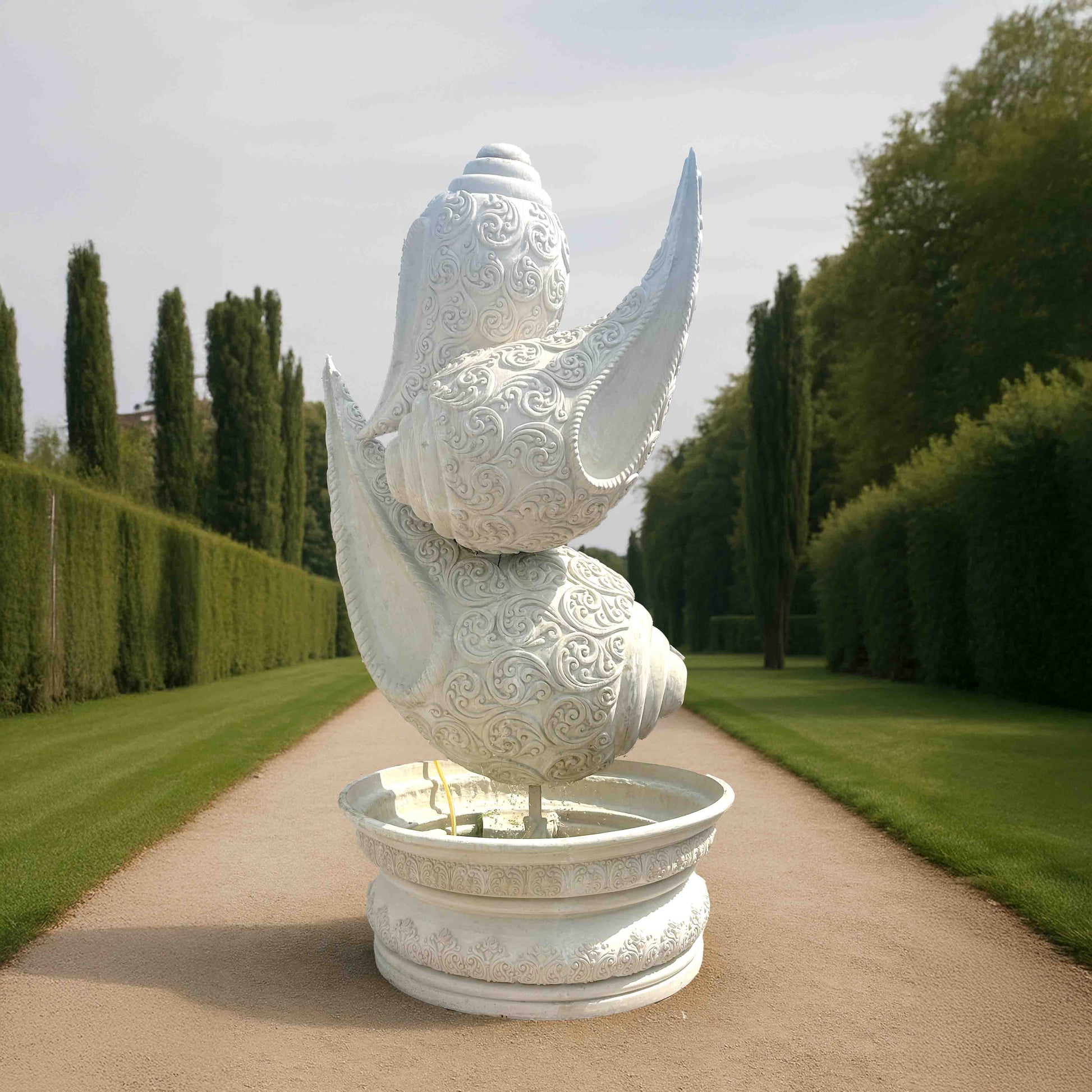 Majestic white marble-finish shankh (conch shell) fountain sculpture with intricate carvings, set on a decorative pedestal basin along a grand garden pathway.