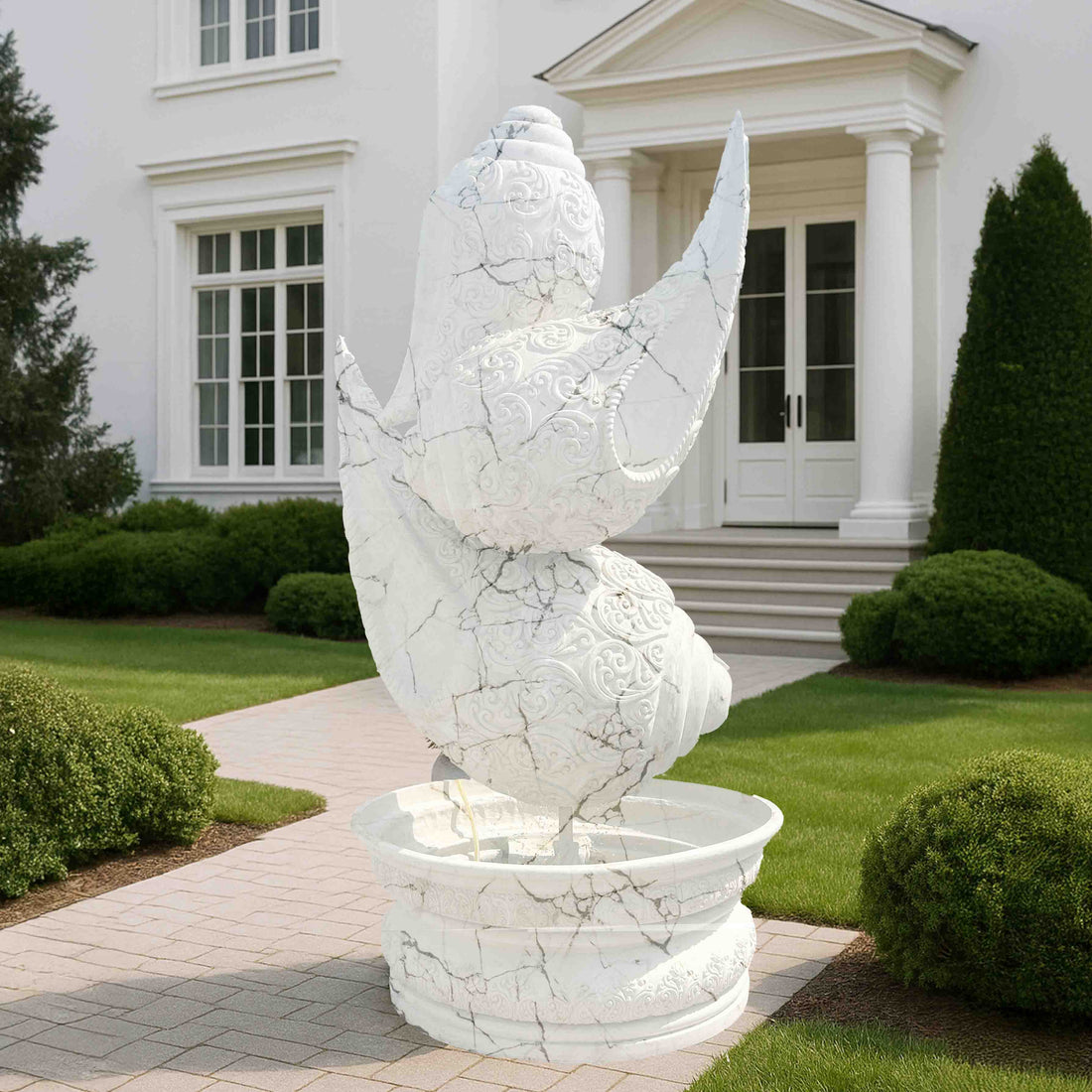Intricately carved white marble-finish shankh (conch shell) fountain sculpture with detailed textures, placed on a pedestal basin in a luxury garden entrance setting.