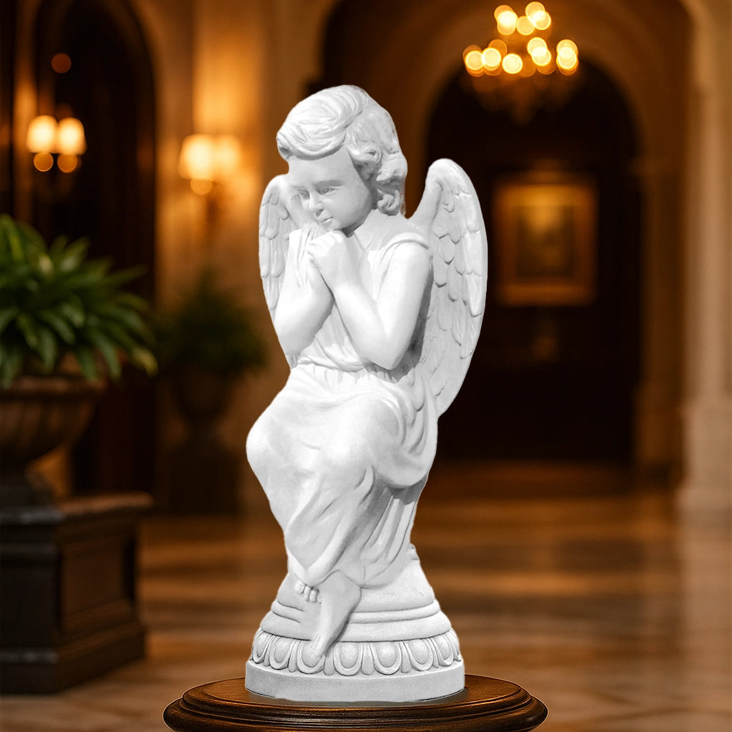 Elegant white praying angel sculpture with wings, crafted in fine detail, placed indoors on a pedestal in a luxury hall with chandelier lighting.
